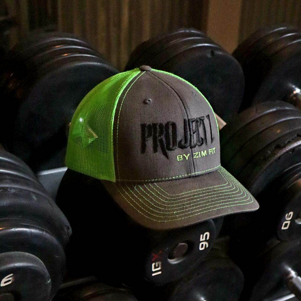A green and brown mesh hat with the 'PROJECT Z' logo in black on the front, placed on a bench with weight plates in the background.